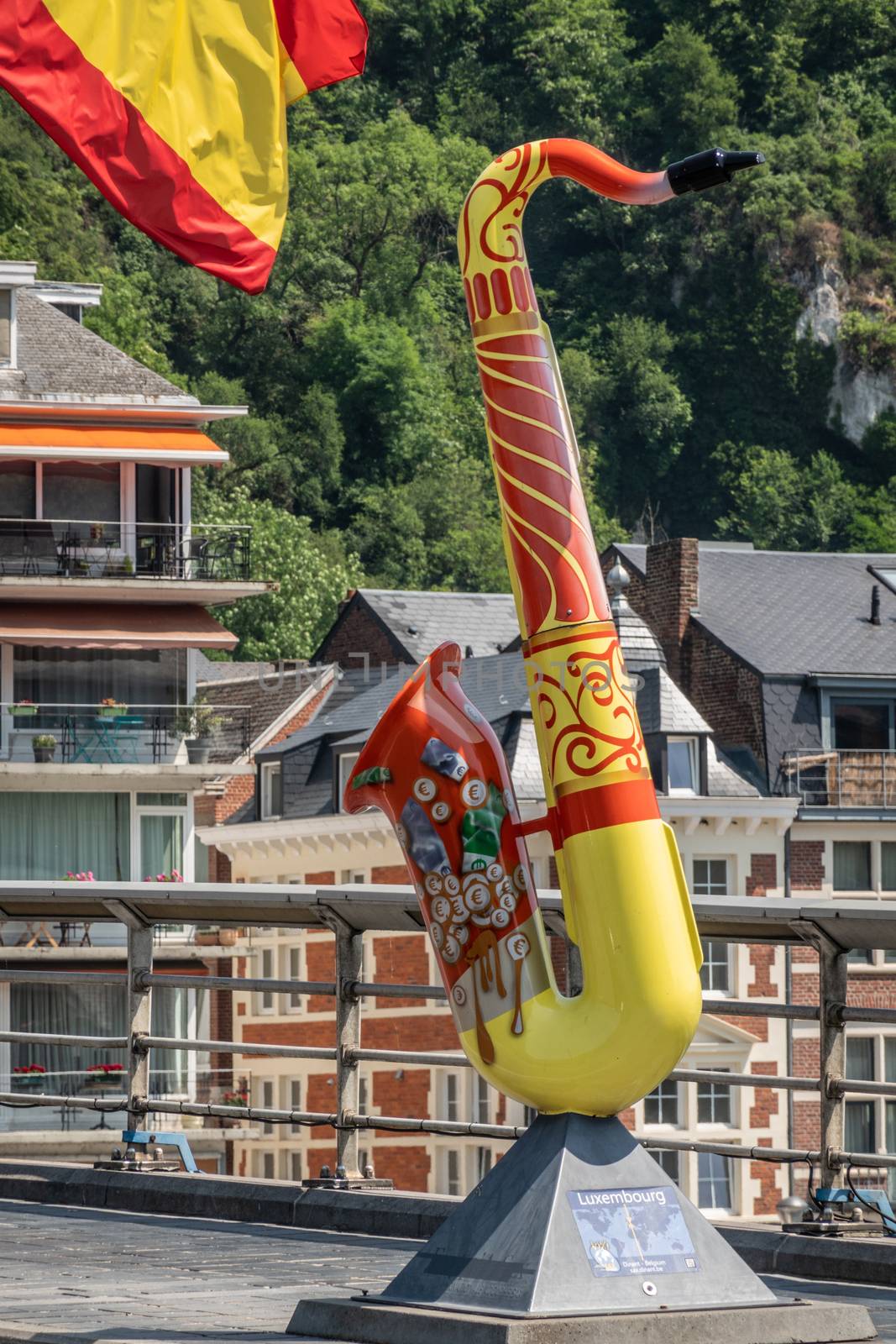 Dinant, Belgium - June 26, 2019: Red and yellow saxophone statue to honor Luxemburg on Charles de Gaulle bridge. Business and houses in back.