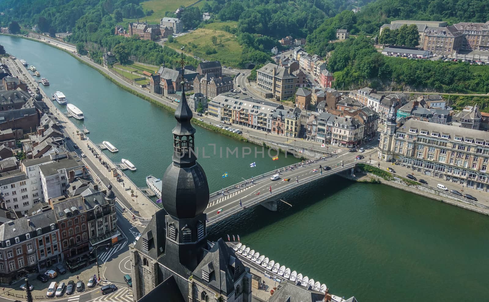 Dinant, Belgium - June 26, 2019: Seen from Citadelle. Charles de Gaulle bridge with focus on south flow of River Meuse. Green landscape, blue sky and plenty of buildings, Church spire.