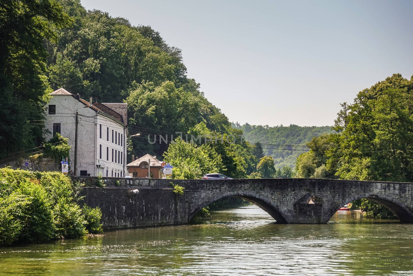 Dinant, Belgium - June 26, 2019: Old roman bridge over greenish Lesse River. Saint Anne Church tower captured in green foliage on the right. White building on the left under light blue sky.