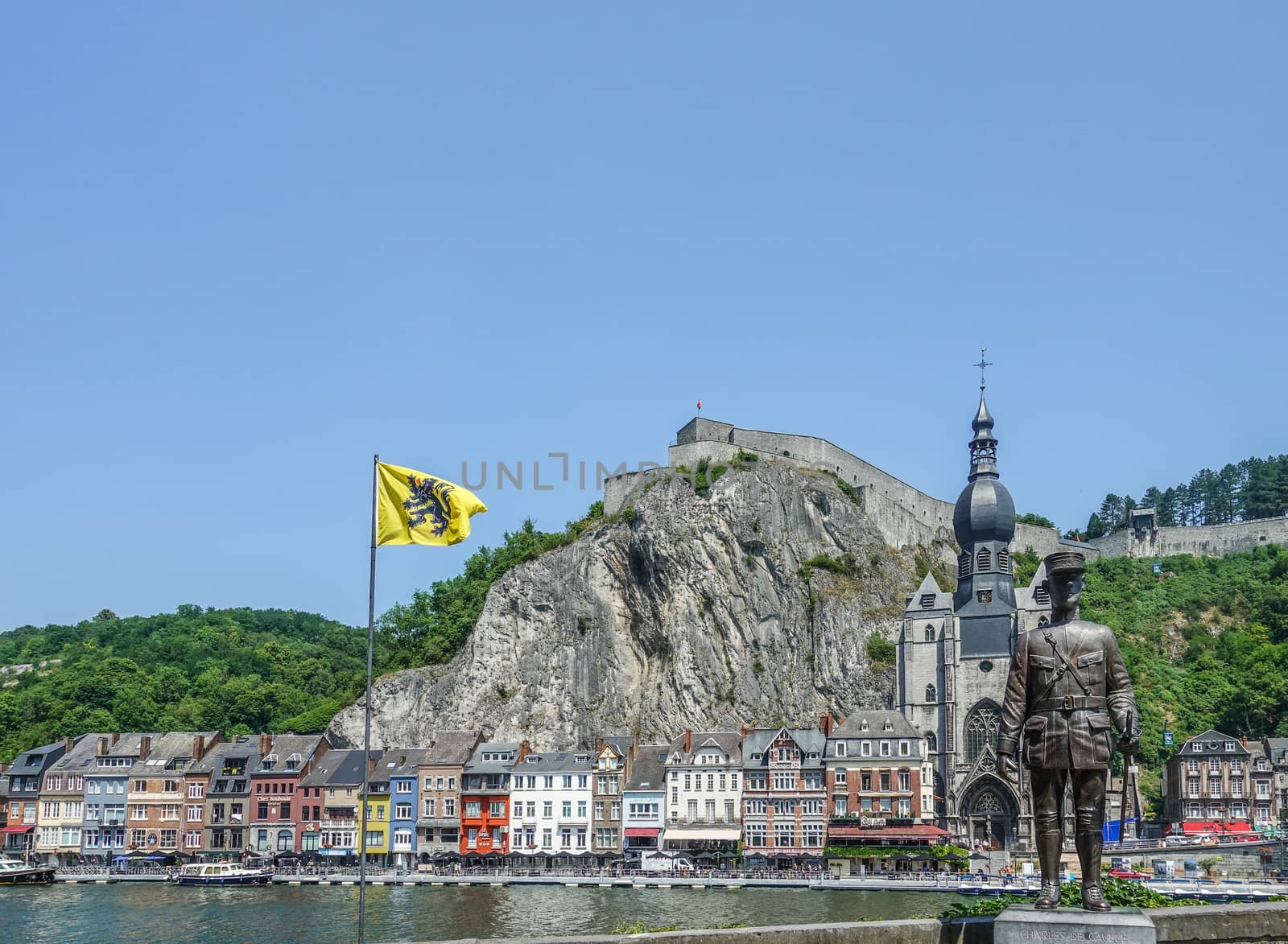 Dinant, Belgium - June 26, 2019: Flag of Flanders, Notre Dame church, Citadelle and Charles De Gaule statue on opposite banks of Meuse River, under blue sky. Green foliage. Multiple flags.