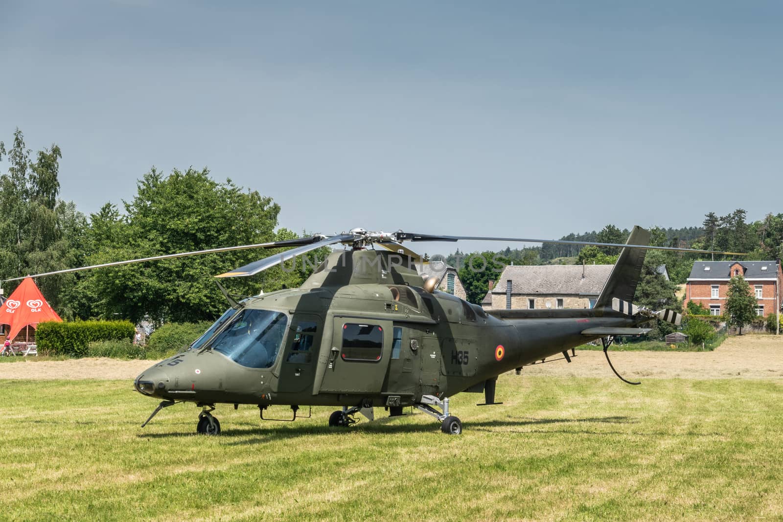 Han-sur-Lesse, Belgium - June 25, 2019: Belgian army green helicopter set on green lawn against foliage and a few houses under blue sky.