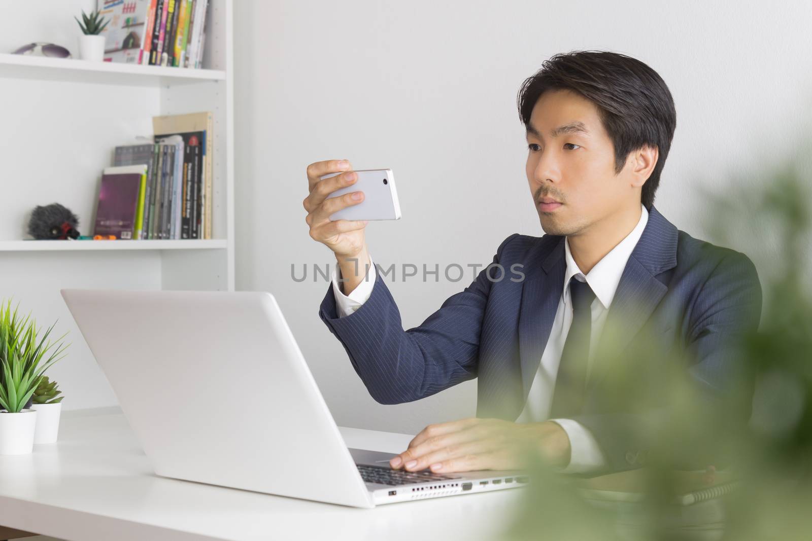 Asian Businessman Take Photo or Selfie by Smartphone in front of Laptop monitor in Office. Asian businessman relax time