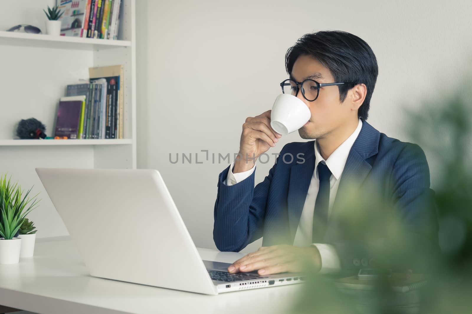 Asian Businessman Wear Eyeglasses and Drink Coffee in front of Laptop. Relax between working time of Asian businessman