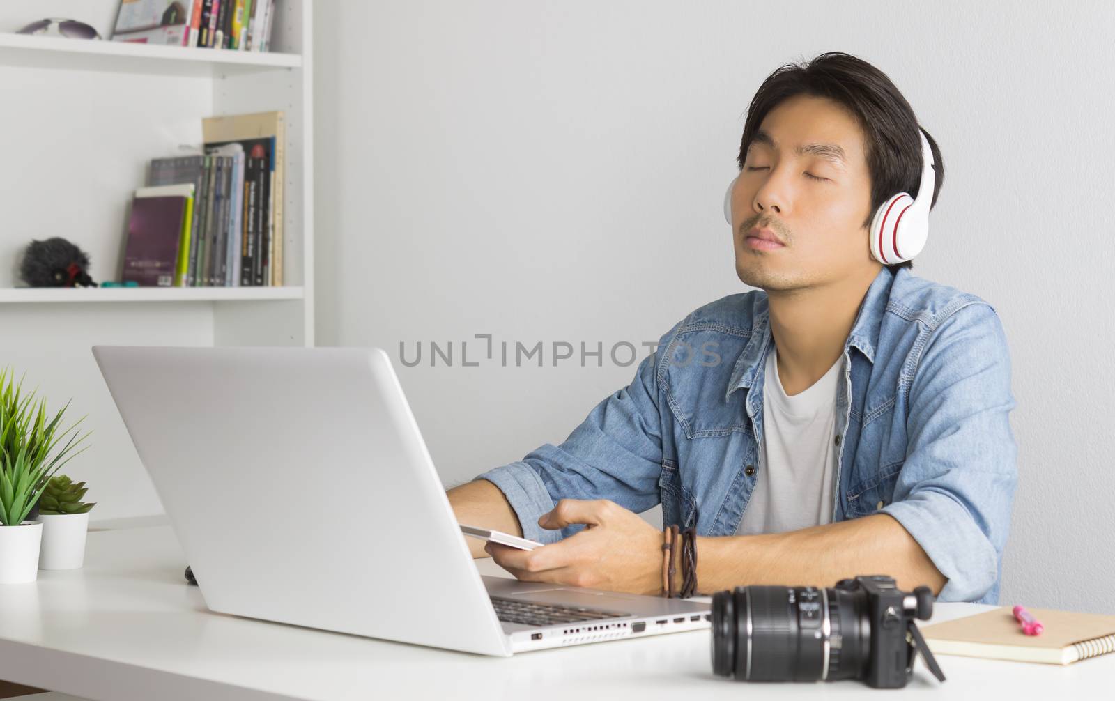 Portrait Asian Photographer with Laptop and Smartphone and Headphone in Home Office. Photographer listening to music by use bluetooth technology in smartphone