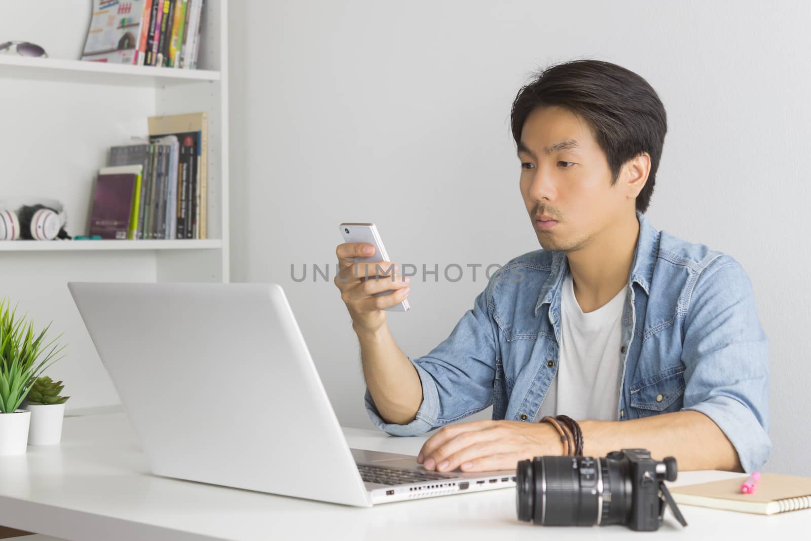 Asian Photographer or Freelancer in Denim or Jeans Shirt Send Message to Customer by Smartphone in front of Laptop in Home Office. Photographer or freelancer working with technology