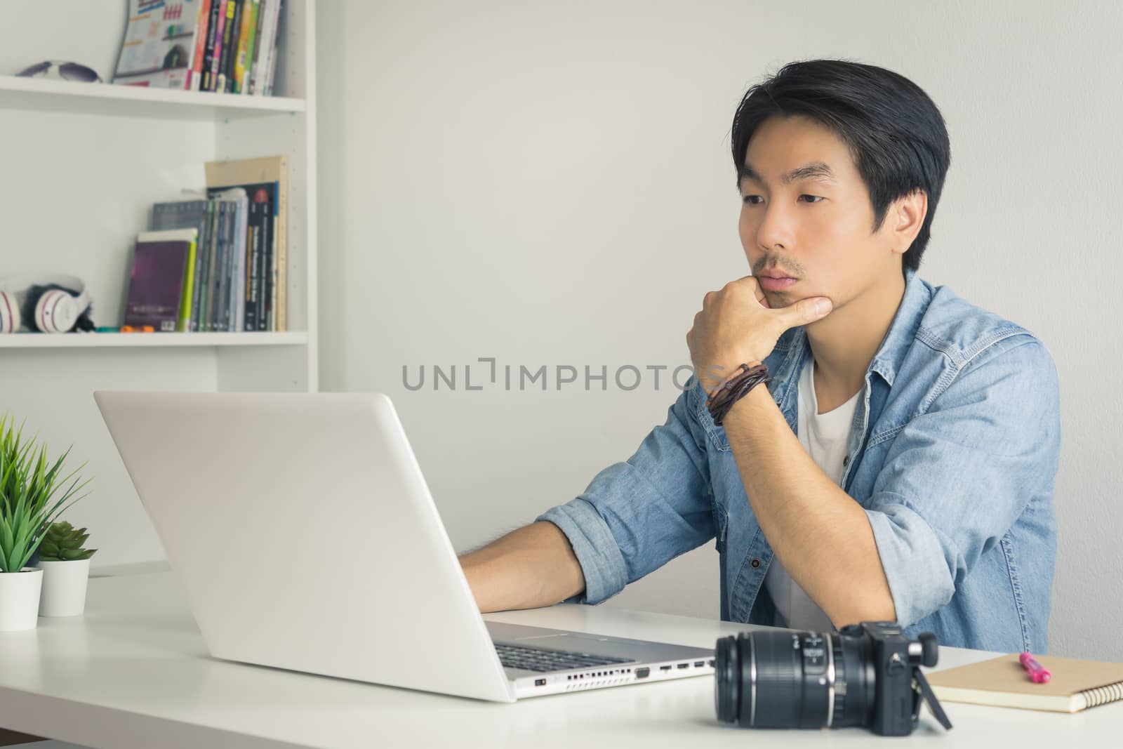 Asian Photographer or Freelancer in Denim or Jeans Shirt Serious Checking Photo File in Laptop in Home Office. Photographer or freelancer working with technology