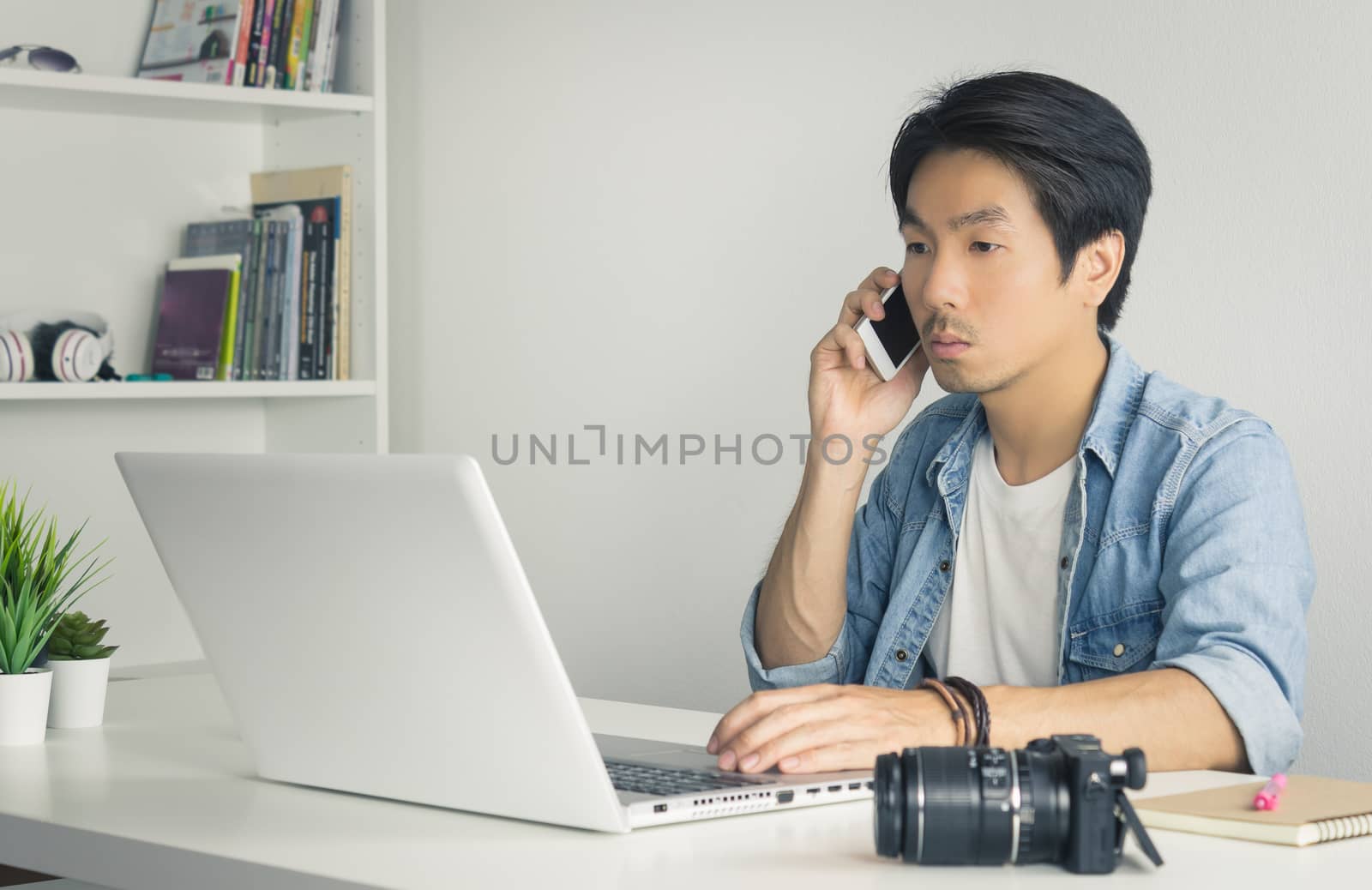 Asian Photographer or Freelancer in Denim or Jeans Shirt Talking with Customer by Smartphone in front of Laptop in Home Office. Photographer or freelancer working with technology