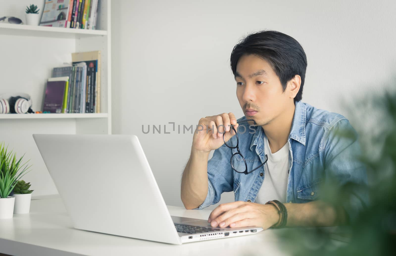 Asian Casual Businessman or Freelancer in Denim or Jeans Shirt Hold Eyeglasses and Working with Laptop in Home Office. Serious casual businessman or Freelancer working with technology