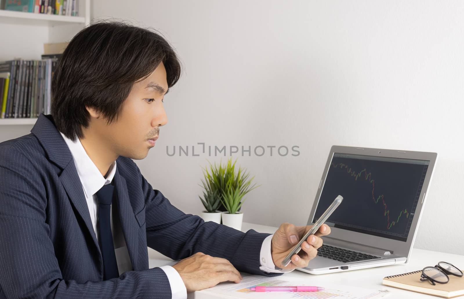 Young Asian Forex Trader or Investor or Businessman in Suit Looking Smartphone and Trading Forex or Stock Chart by Laptop in Trader Room