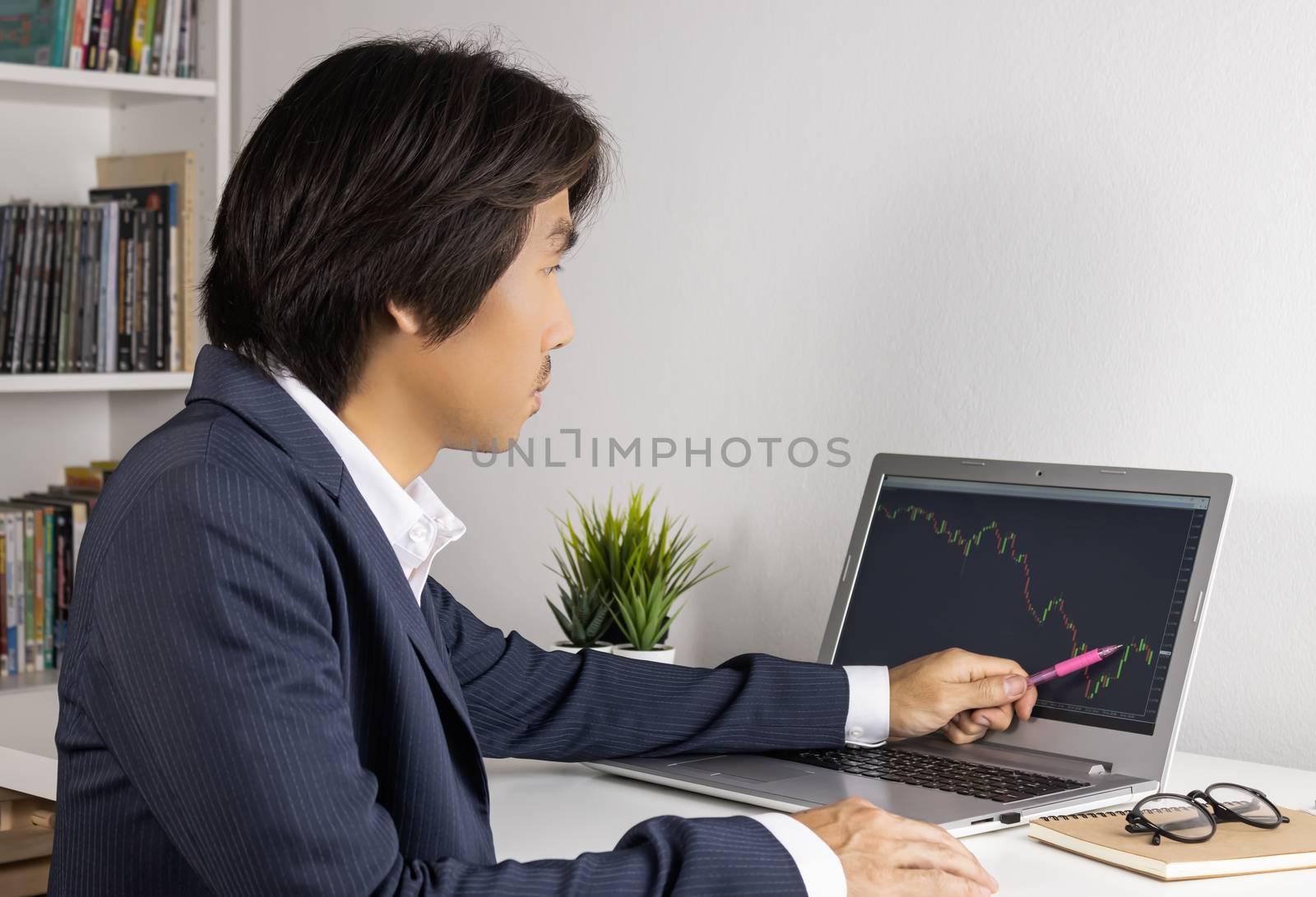 Young Asian Forex Trader or Investor or Businessman in Suit Trading and Pointing Forex or Stock Chart on Laptop Screen in Trader Room