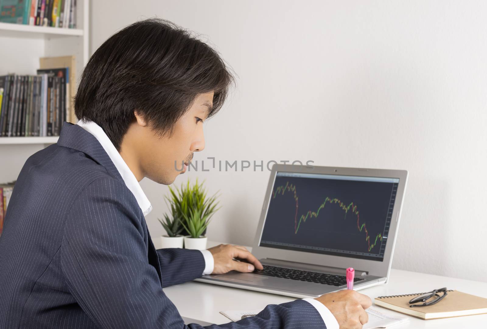 Young Asian Forex Trader or Investor or Businessman in Suit Writing Financial Report and Trading Forex or Stock in Laptop Monitor in Trader Room