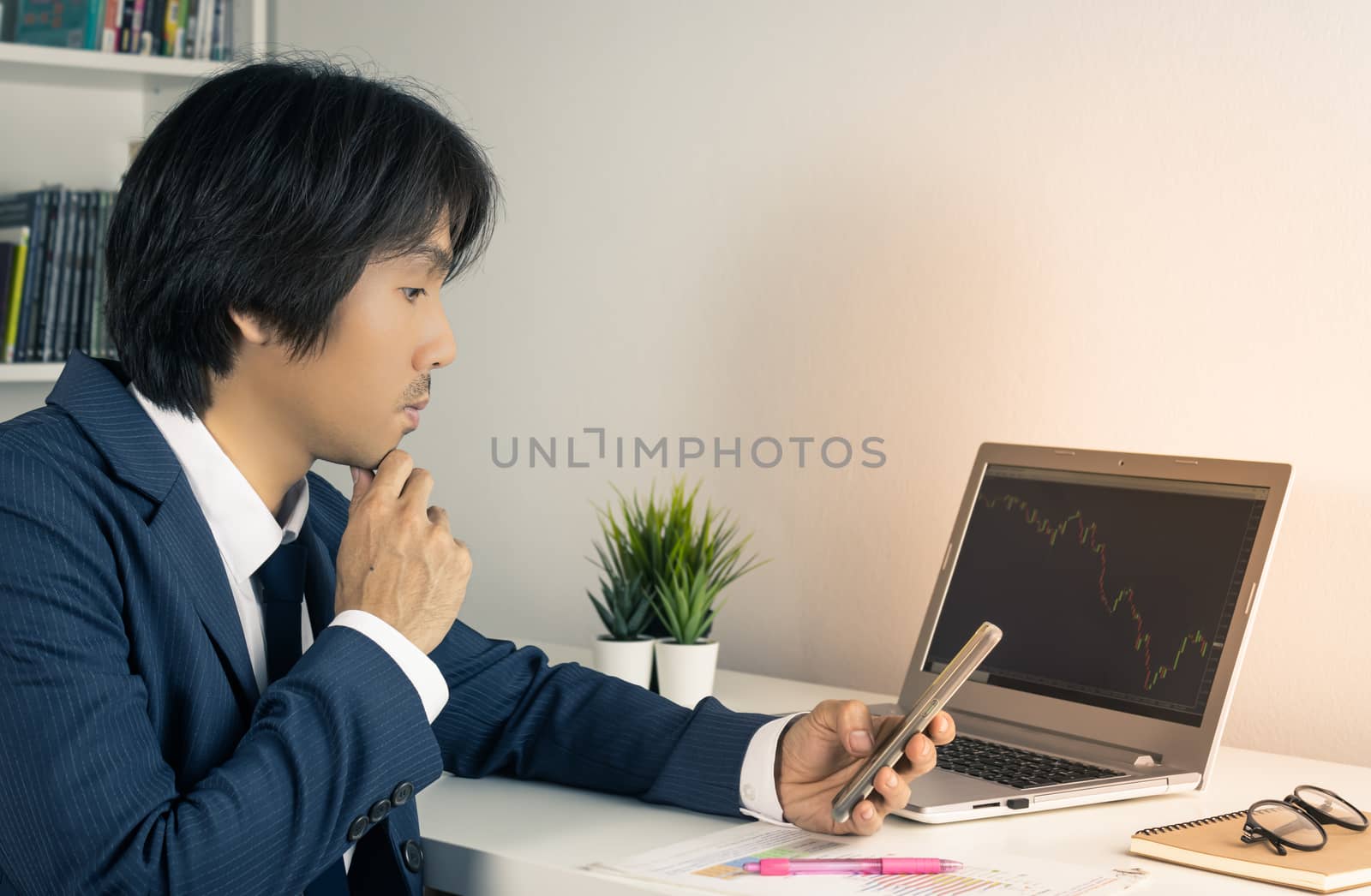 Serious Young Asian Forex Trader or Investor or Businessman in Suit Hold Smartphone and Trading Forex or Stock Chart by Laptop in Trader Room in Vintage Tone