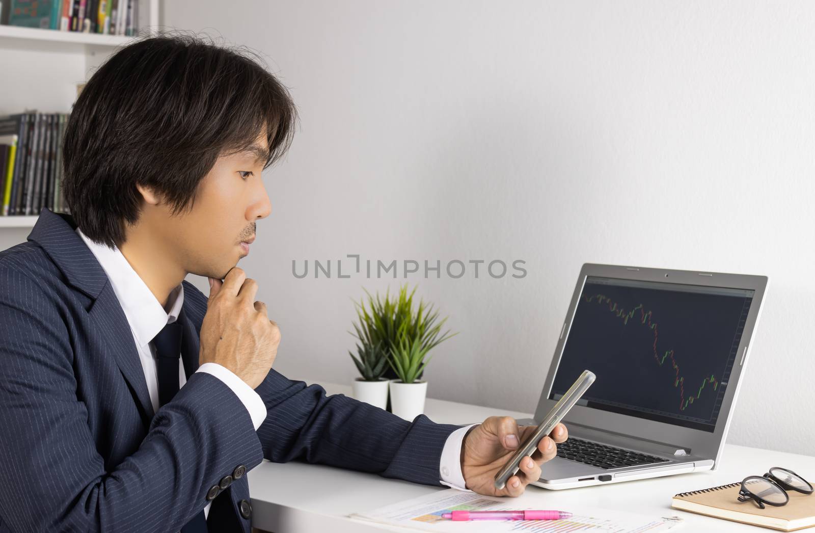 Serious Young Asian Forex Trader or Investor or Businessman in Suit Hold Smartphone and Trading Forex or Stock Chart by Laptop in Trader Room