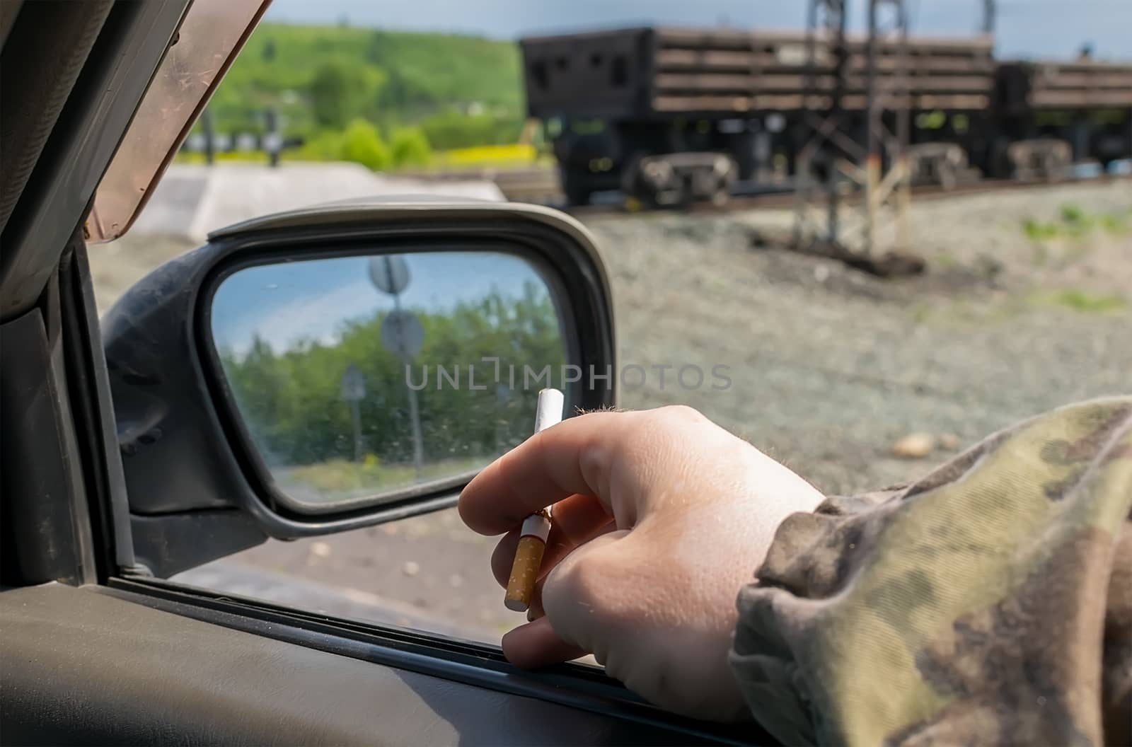 hand men, military, in camouflage clothing, holding a cigarette while in the car, which is near the railway crossing with freight cars