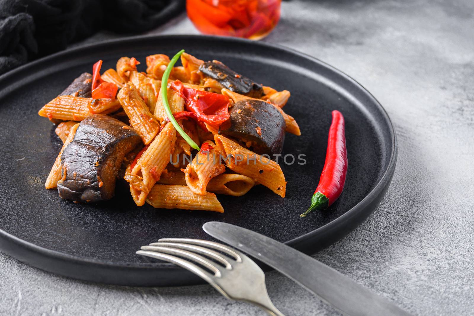 Aubergine penne eggplant pasta, pepper tomatoe sauce, on black plate overgrey concrete background served side view