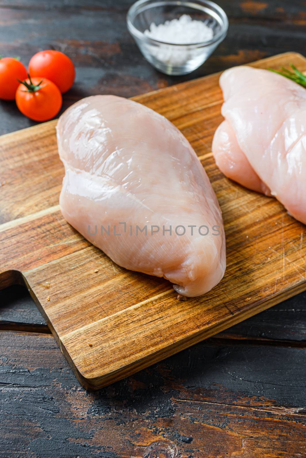 Raw chicken breasts and spices on wooden cutting board on old dark table side view close up.