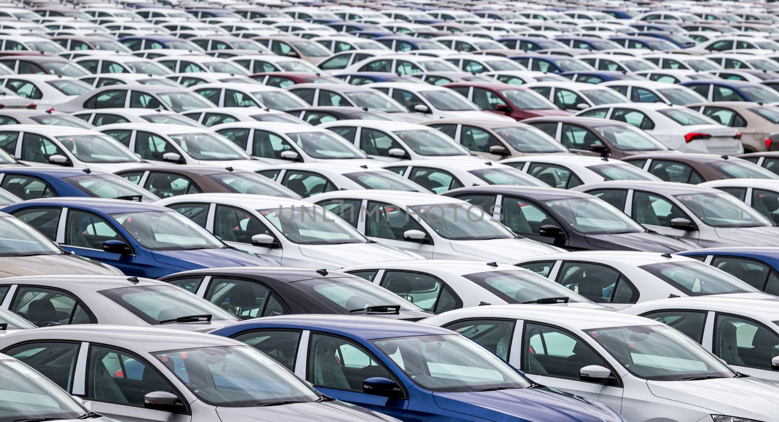 Rows of a new cars parked in a distribution center on a car factory on a cloudy day. Top view to the parking in the open air. by Eugene_Yemelyanov