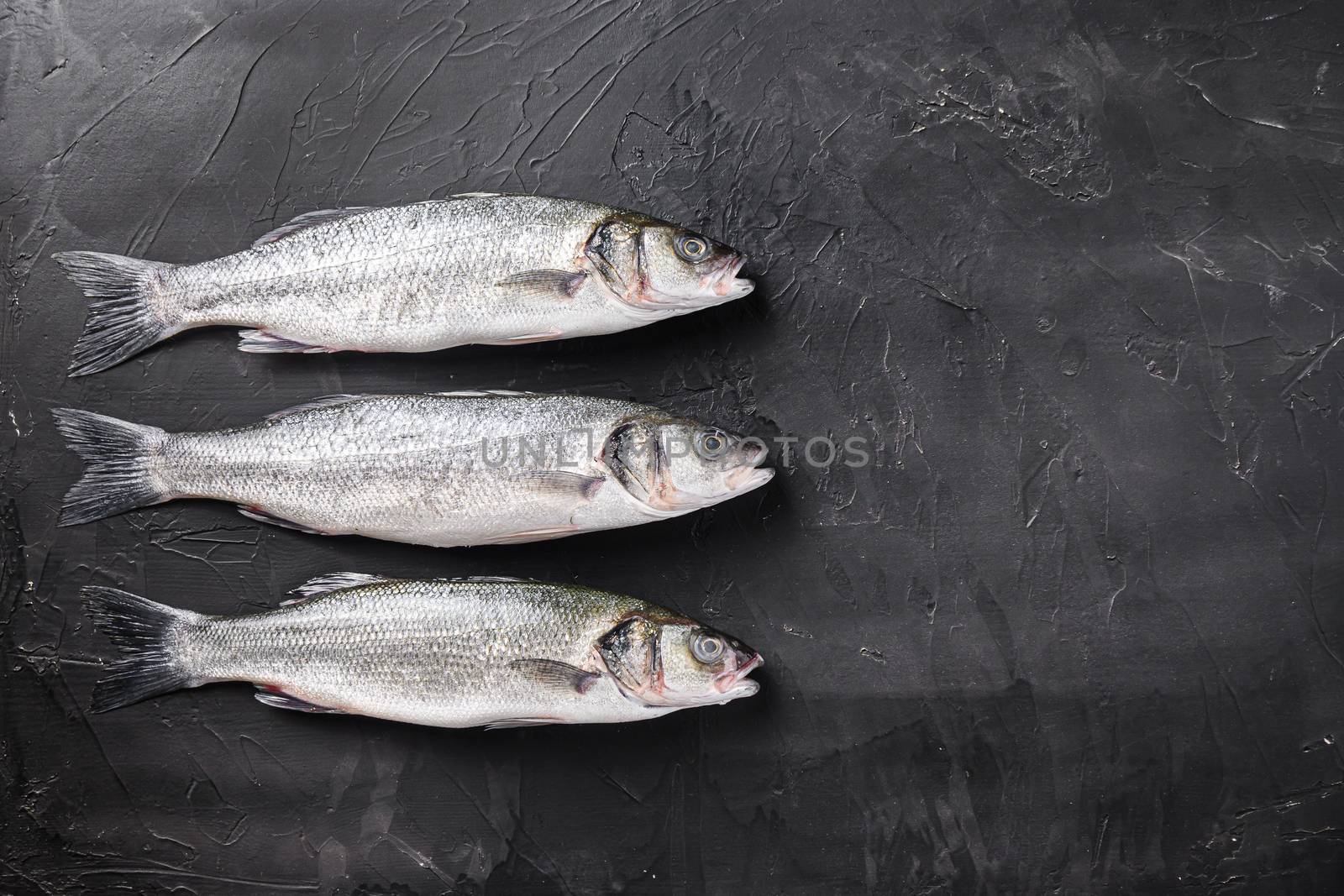 Set of whole raw sea bass with spices and herbs ingredients on chopping board over black stone table top view space for text