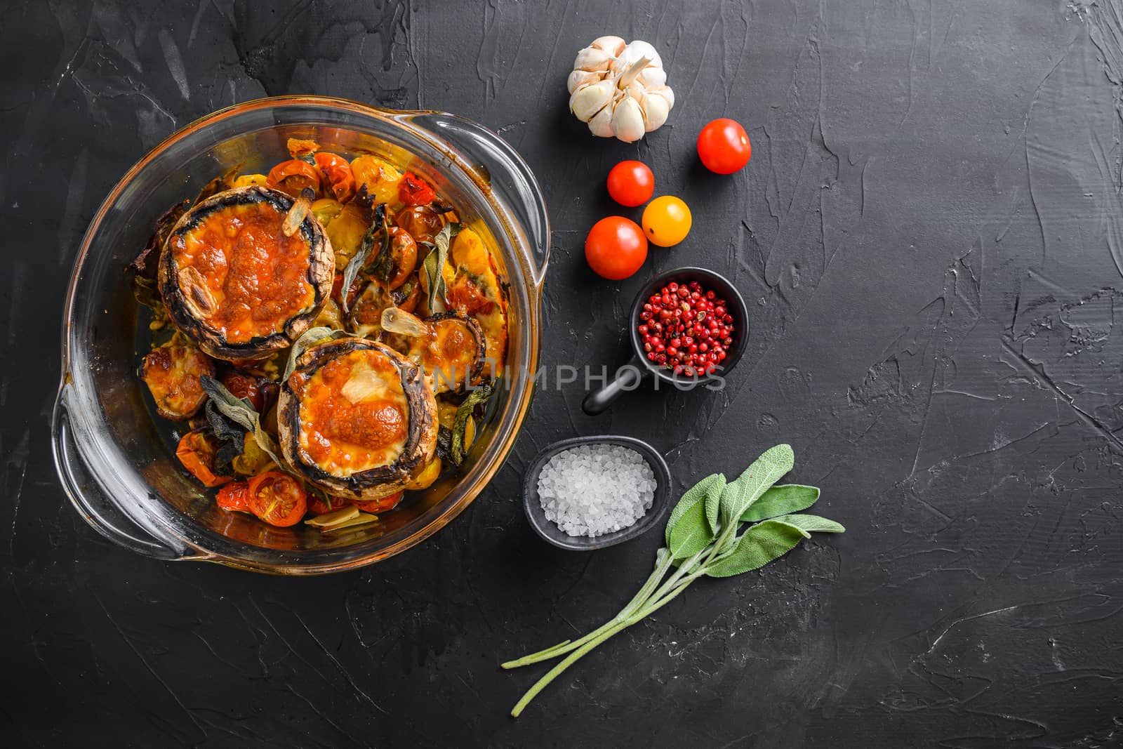 Portobello mushrooms,baked with ingredients cheddar cheese, cherry tomatoes and sage in glass pot on black stone background top view space for text