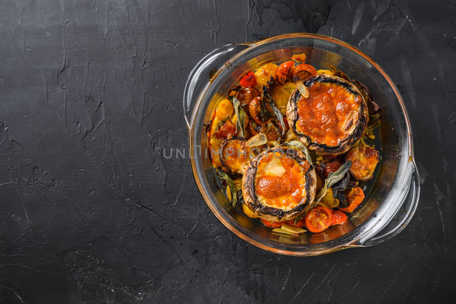 Portobello mushrooms,baked with cheddar cheese, cherry tomatoes and sage in glass pot on black stone background top view space for text