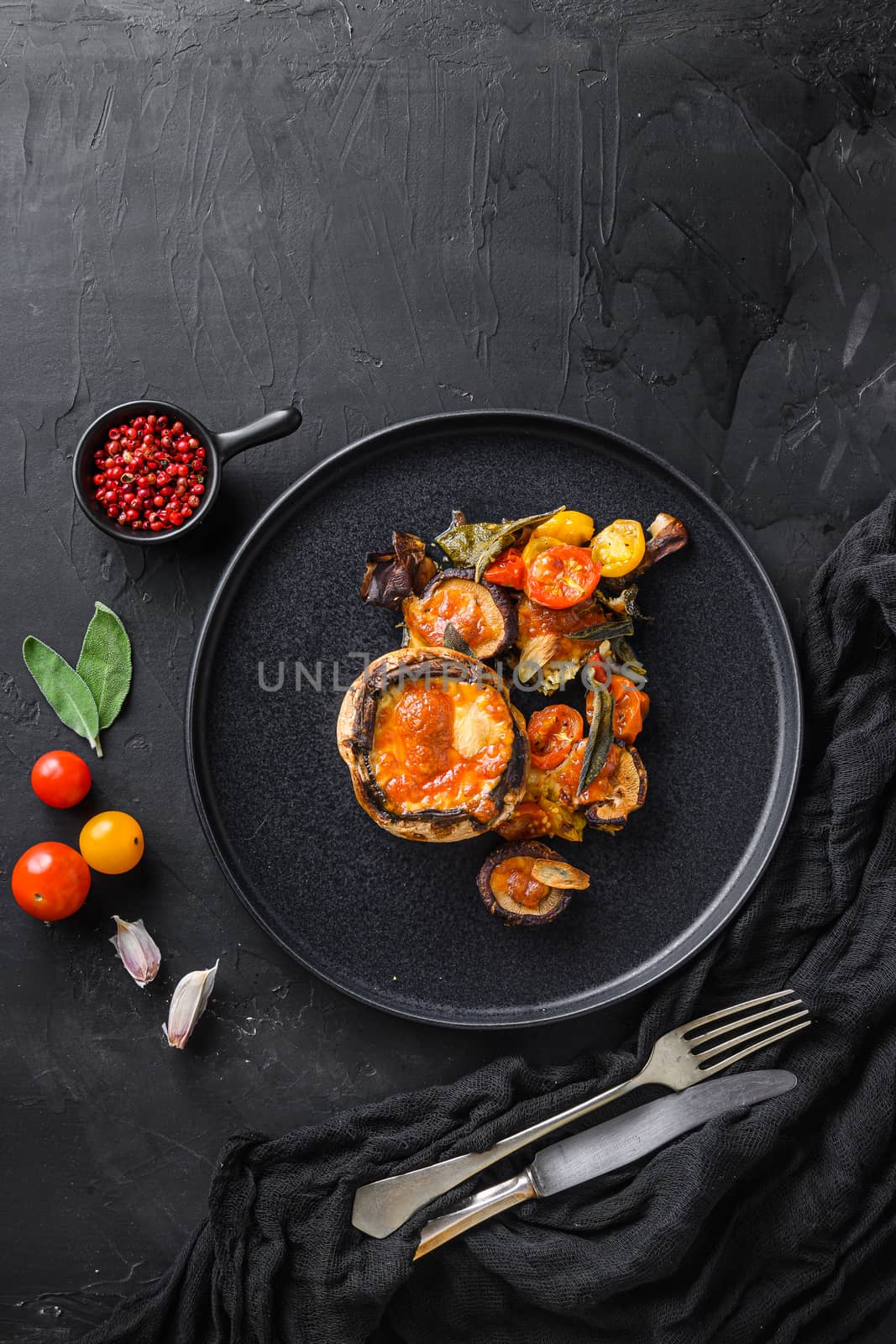 Served portobello mushrooms,baked and stuffed with cheddar cheese, cherry tomatoes and sage on black plate over black background top view space for text