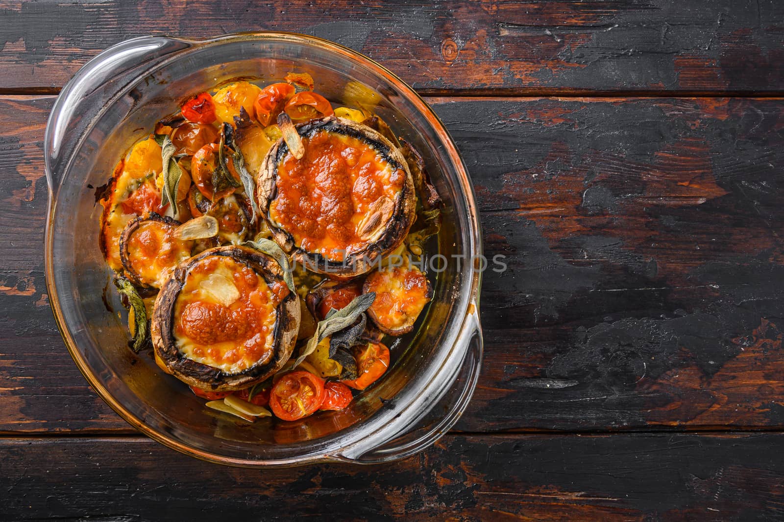 Portobello baked with cheddar cheese, cherry tomatoes and sage in glass pot on old wooden background top view space for recipe or text