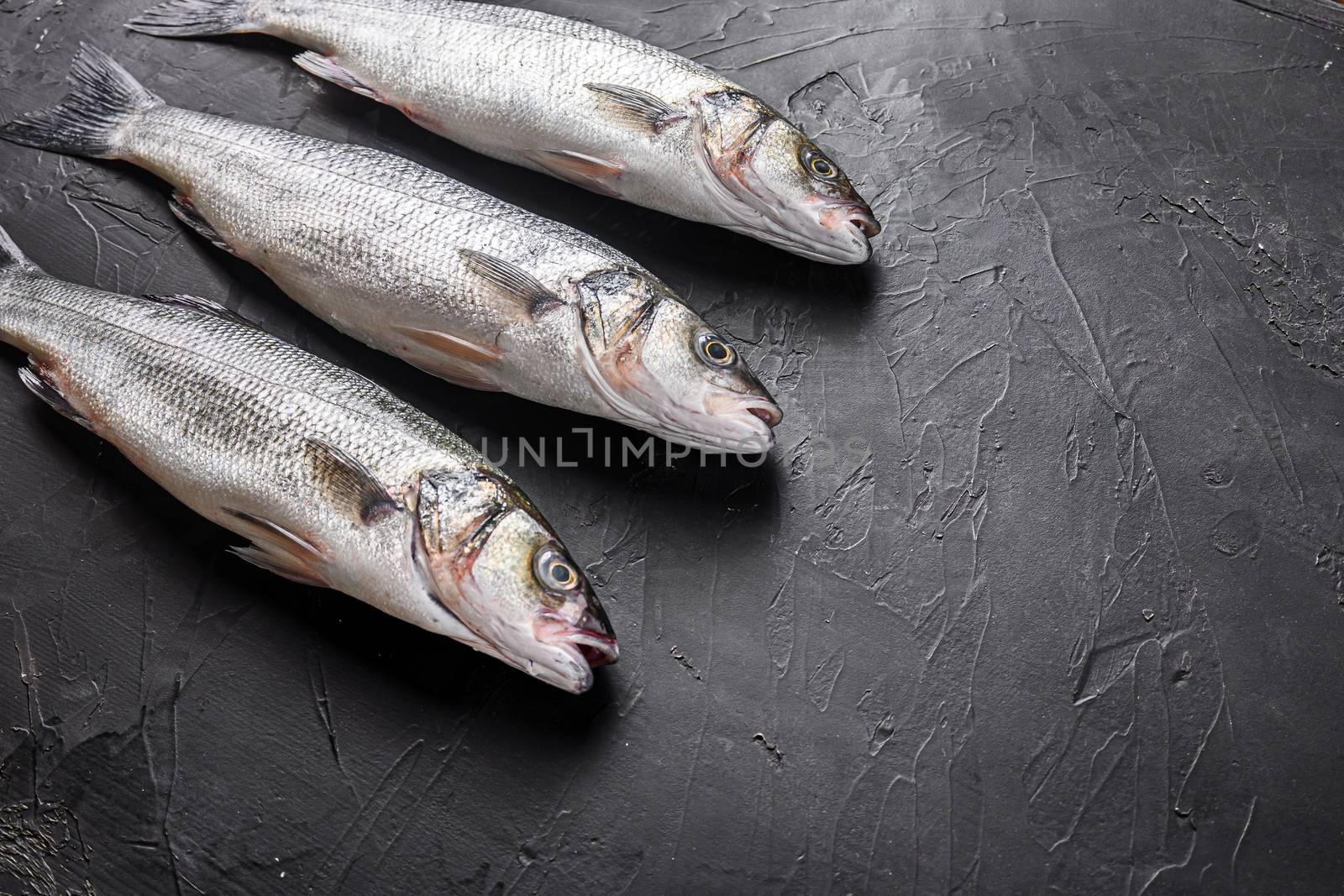 Set of whole raw seabass with spices and herbs ingredients on chopping board over black stone table side view