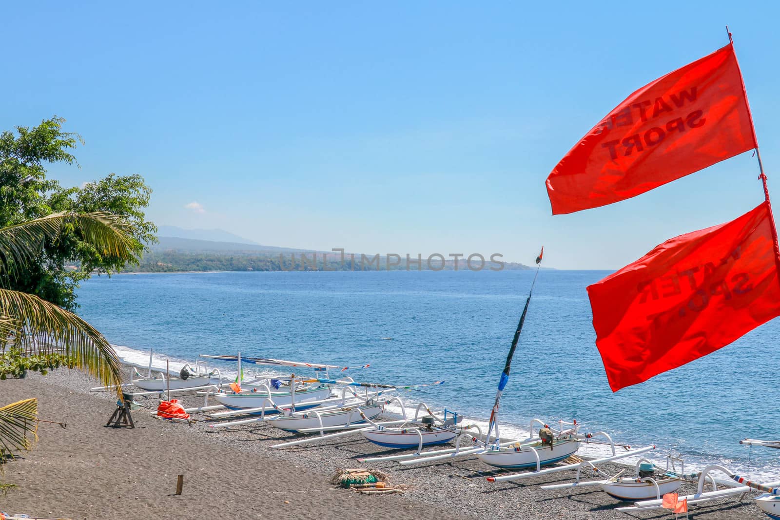 Black volcanic sand beach from nearby Agung volcano. Tropical paradise with palm trees and majestic volcano in the background. Red flag in the wind.