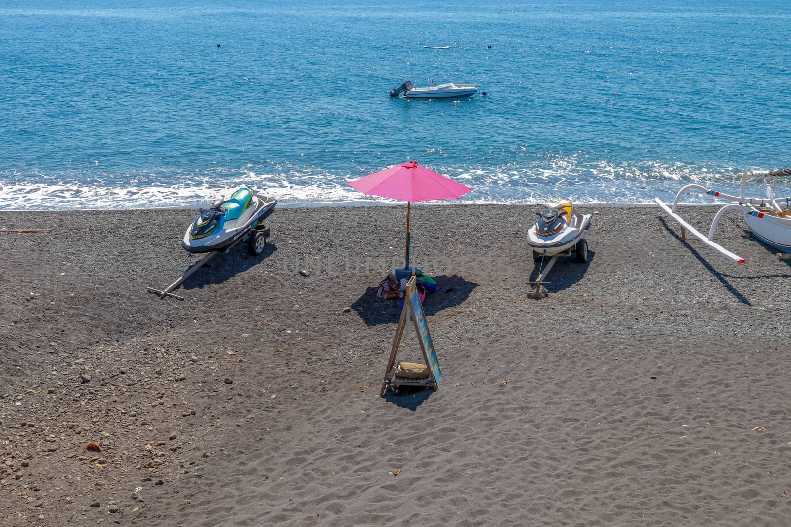 Jetski Seadoo parked on the beach. Black volcanic sand beach. In the background of the sea and moored boat. Red parasol.