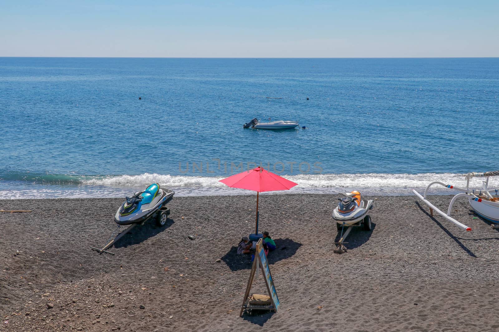 Jetski Seadoo parked on the beach. Black volcanic sand beach. In the background of the sea and moored boat. Red parasol.