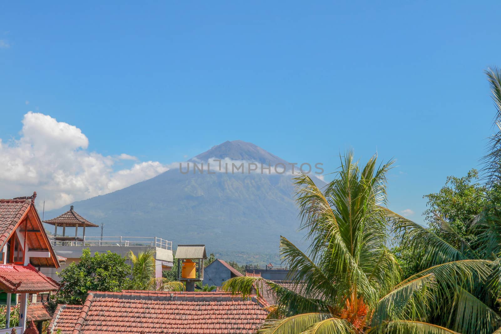 Tropical paradise with palm trees and majestic volcano in the background. White clouds around a volcano crater. View from above the roofs of the house.