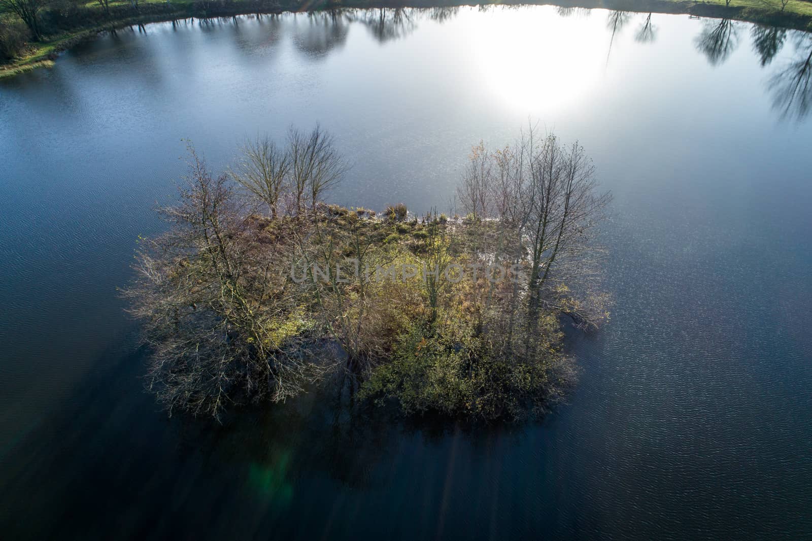 Small island in a pond, backlit with the reflections of the sun in the water, aerial view with drone