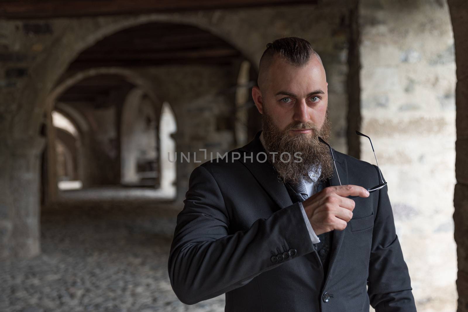 Man with short hair and long beard holding sunglasses while posing in the arcades of an ancient village, portrait set