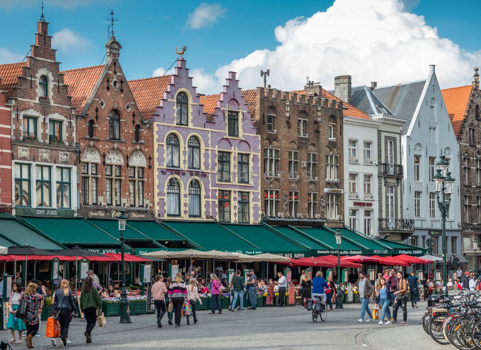 Bruges, Flanders, Belgium -  June 15, 2019: Brick stone facade row with step gables, now restaurants and bars with colored awnings, on NW side of Markt Square. People walking.