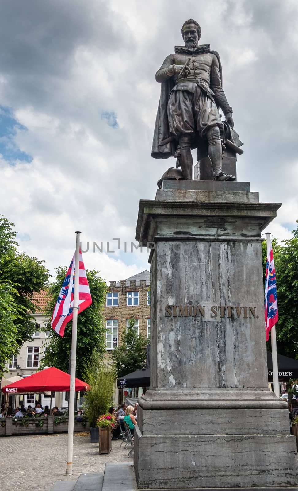 Bruges, Flanders, Belgium -  June 17, 2019: Simon Stevin statue on his own square where people enjoy drinks under cloudscape. Flags and green foliage.
