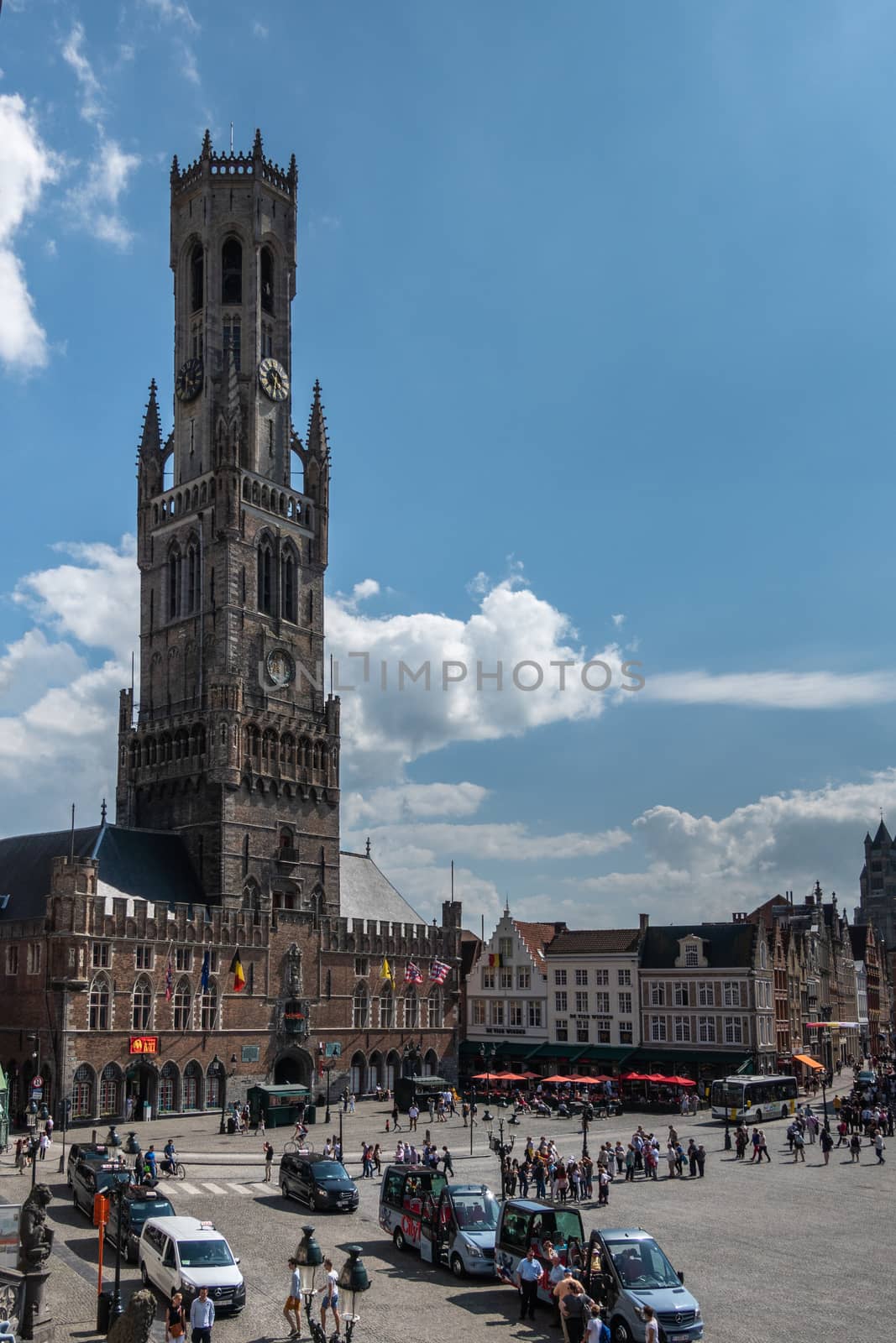 Bruges, Flanders, Belgium -  June 17, 2019: Belfry and tall clock tower, Halletoren, behind Markt square with people, taxis and tourist buses. All under blue sky with white clouds.