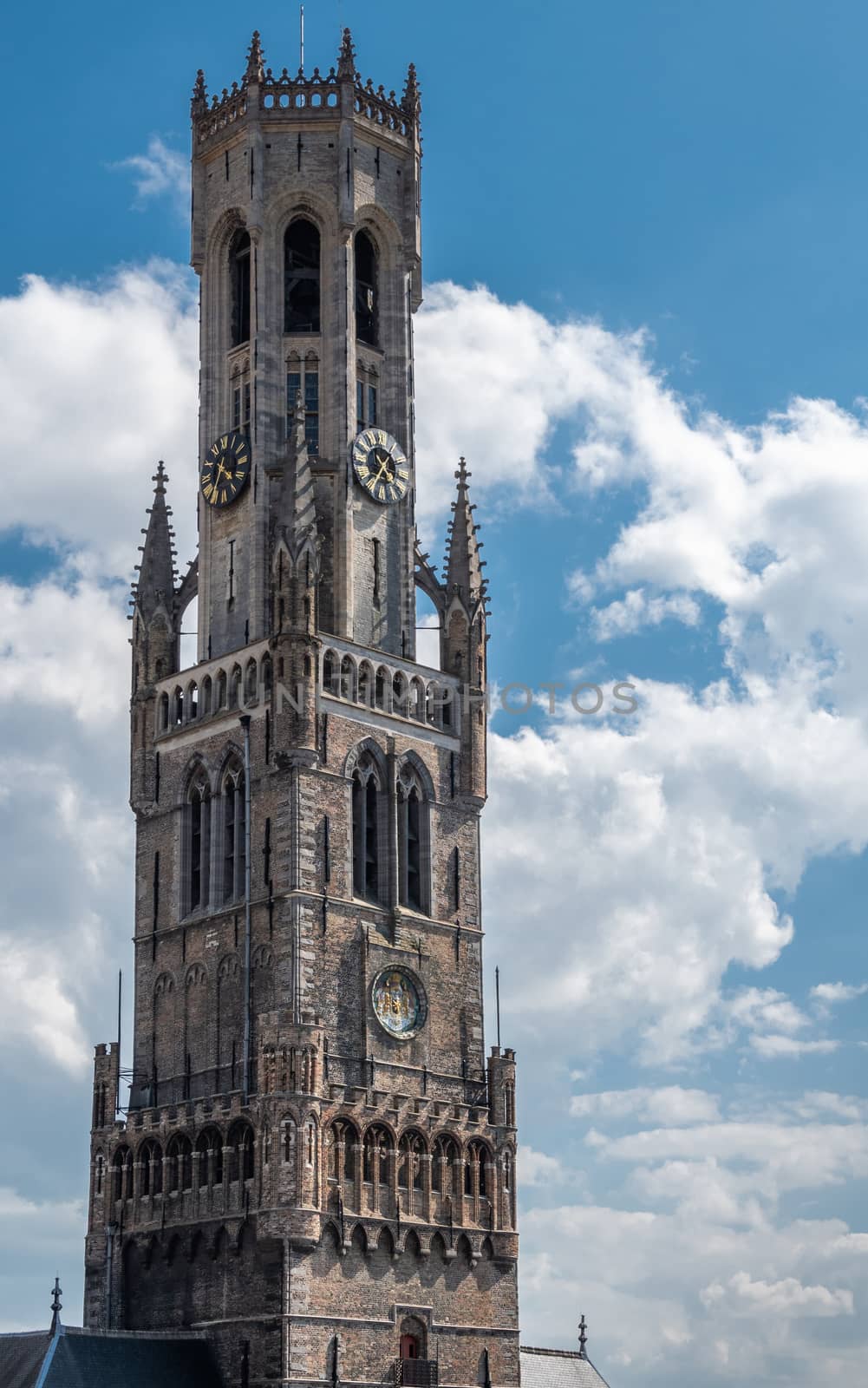 Bruges, Flanders, Belgium -  June 17, 2019: Closeup of Belfry tall gray brick clock tower, Halletoren, against blue sky with white clouds.