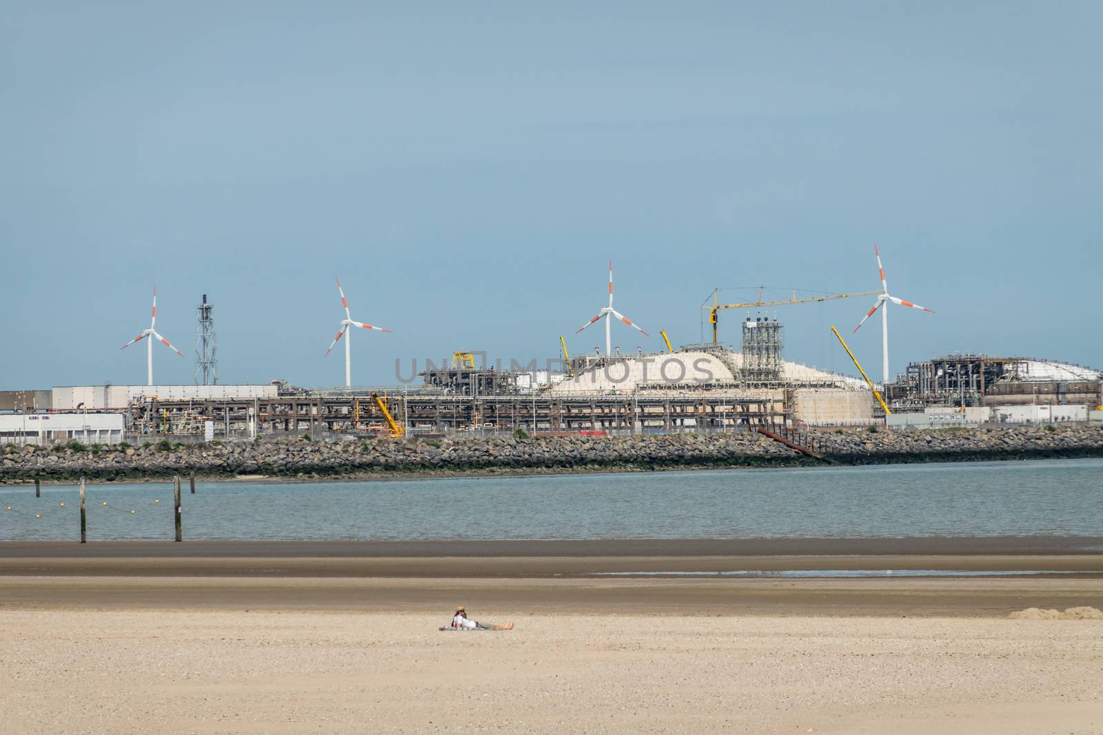 Zeebrugge, Flanders, Belgium -  June 18, 2019: Closeup of LNG terminal in port of Zeebrugge under blue sky as seen from beach in Knokke-Heist. Lone sun bather on sand.