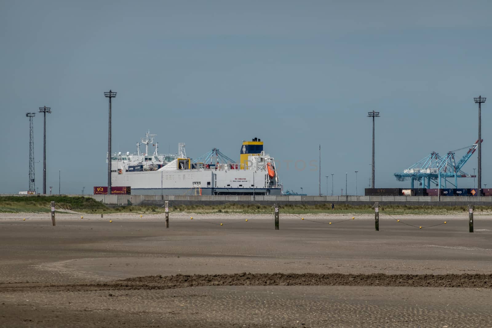 Zeebrugge, Flanders, Belgium -  June 18, 2019: Ferry and container ship with Container cranes in port of Zeebrugge, as seen from beach in Knokke-Heist. Gray-blue sky, wet sand.