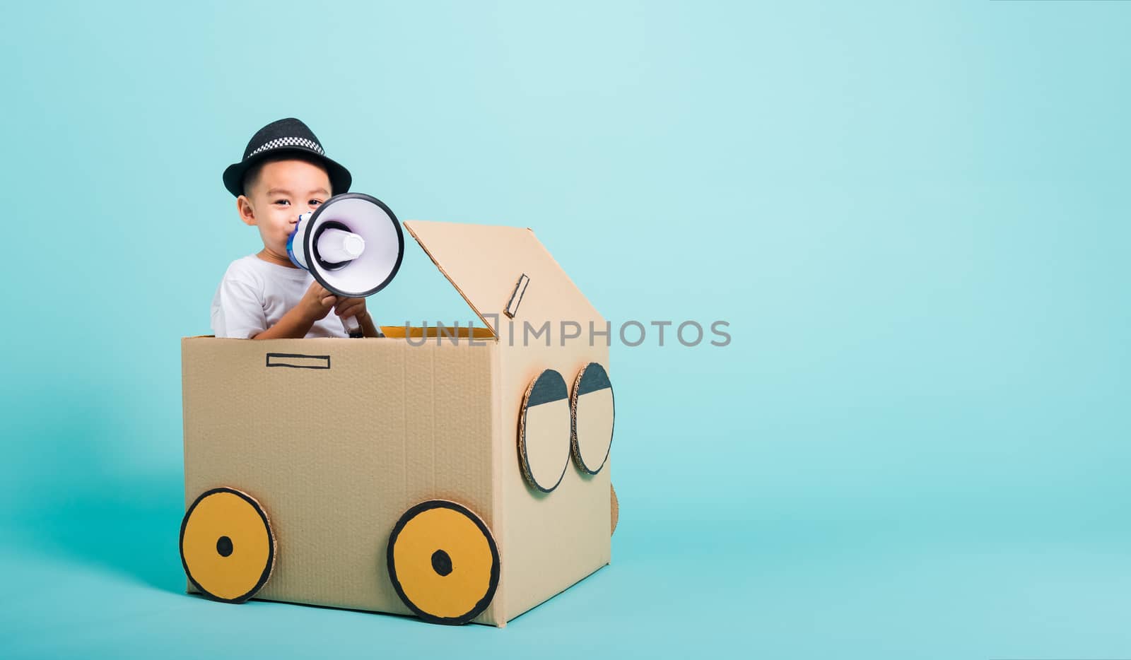 Happy Asian children boy smile in driving play car creative by a cardboard box imagination with megaphone, summer holiday travel concept, studio shot on blue background with copy space for text