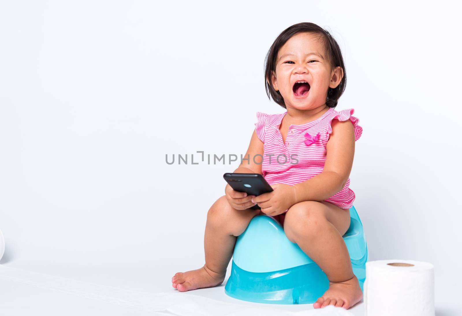Asian little cute baby child girl education training to sitting on blue chamber pot or potty and play smart mobile phone with toilet paper rolls, studio shot isolated on white background, wc toilet