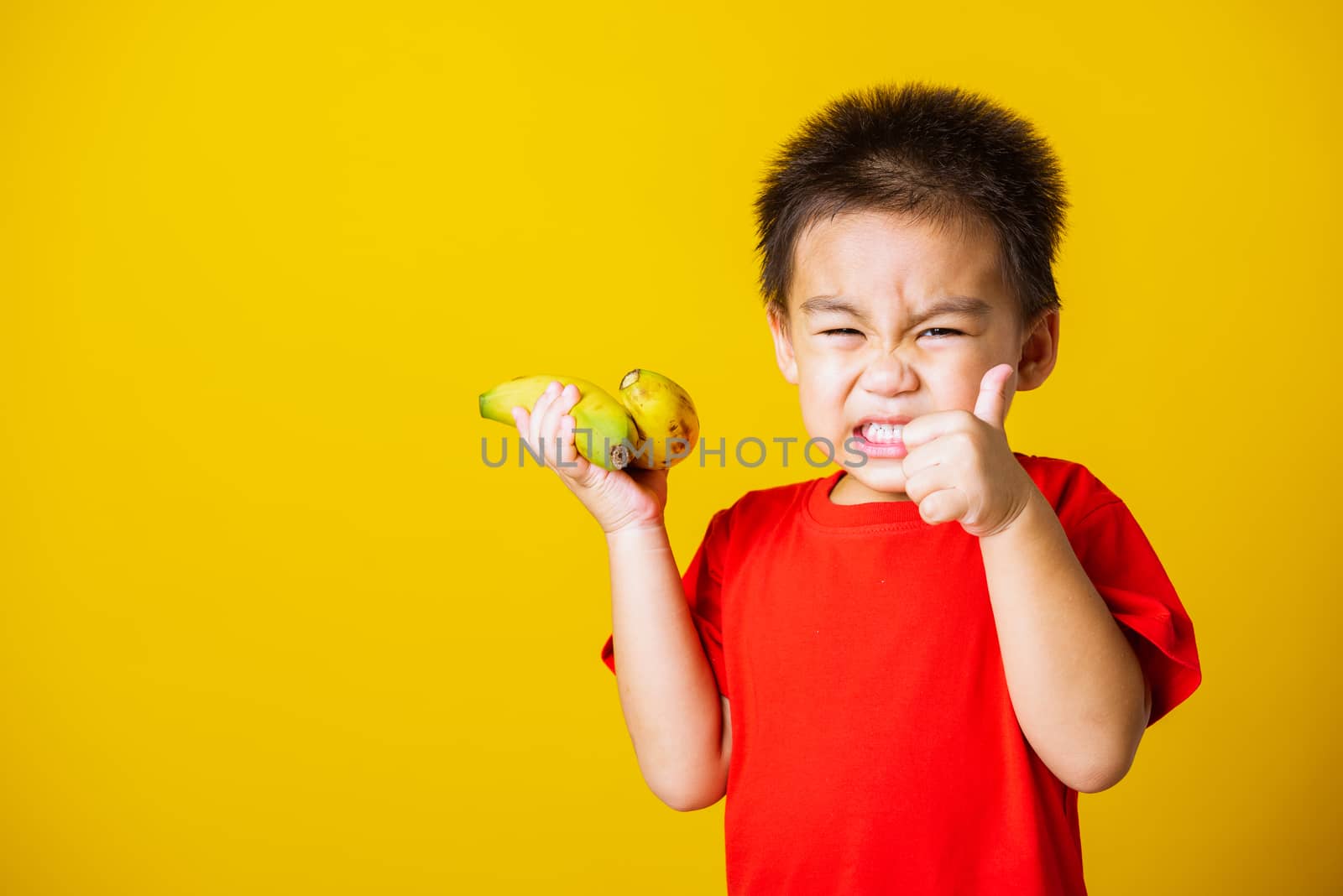 Happy portrait Asian child or kid cute little boy attractive smile wearing red t-shirt playing holds bananas and show finger thumb for good sign, studio shot isolated on yellow background