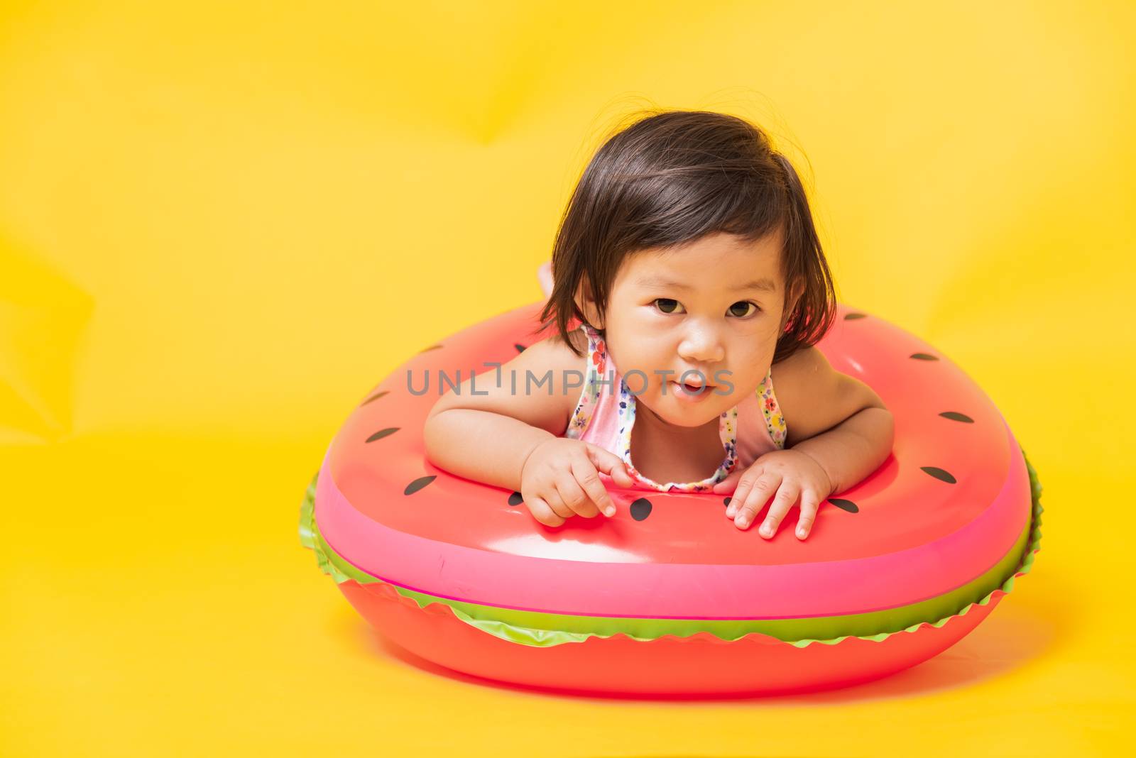 Summer vacation concept, Portrait Asian happy cute little baby girl wear swimsuit sitting in watermelon inflatable ring, Kid have fun sit in inflatable, studio shot isolated yellow background