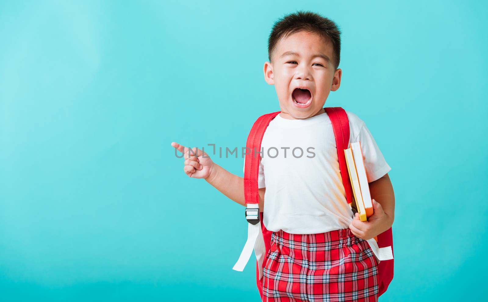 Back to school concept. Portrait Asian happy funny cute little child boy smile hug books and point finger to side away space, isolated blue background. Kid from preschool kindergarten with school bag