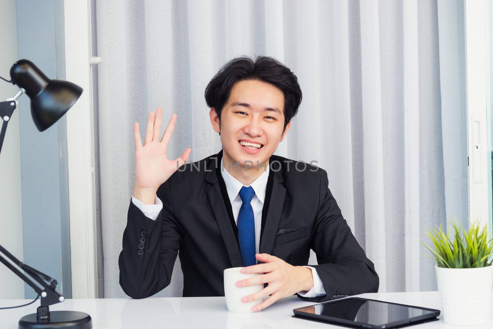 Work from home, Asian young businessman video conference call or facetime he smiling looking to camera sitting on desk using smart digital tablet computer raise hand to say hello at home office