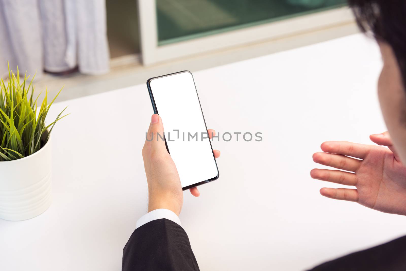 Work from home, Back view of Asian young businessman smile wearing suit video conference call or facetime by smart digital mobile phone blank screen raise his hand to explain to teammates on desk