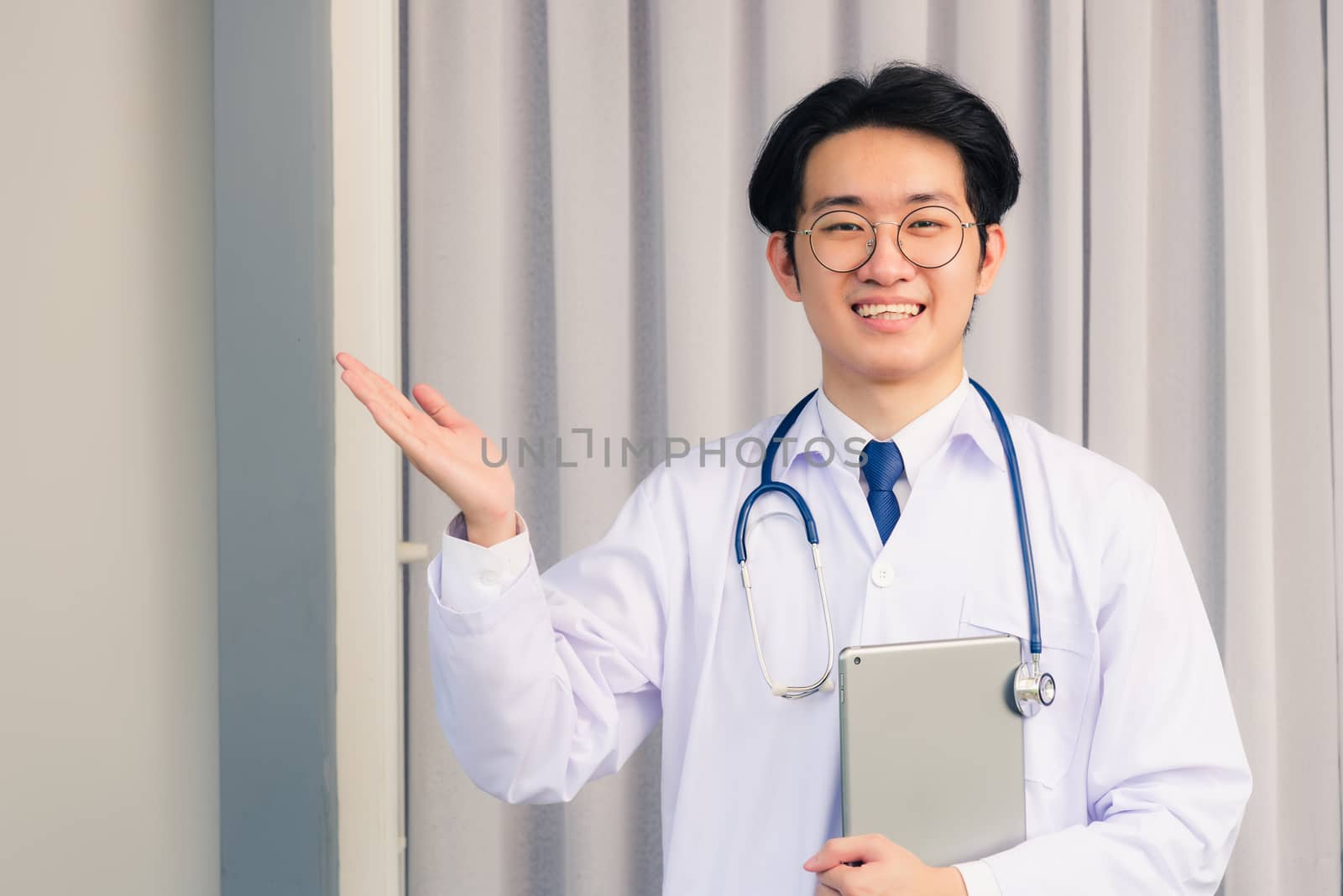 Portrait closeup of Happy Asian young doctor handsome man smiling in uniform and stethoscope neck strap holding smart digital tablet on hand and showing hand to side away, healthcare medicine concept