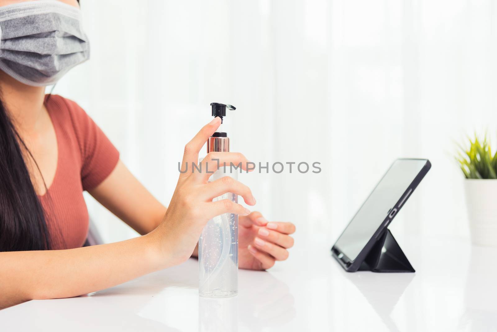 Asian Business young woman working from home office he quarantines disease coronavirus or COVID-19 wearing a protective mask and cleaning hands with sanitizer gel on front tablet computer