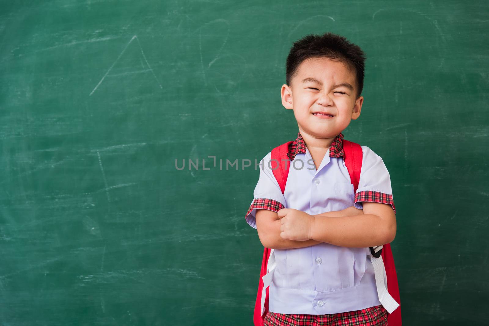 Back to School. Happy Asian funny cute little child boy from kindergarten in student uniform with school bag stand smile crossed arm on green school blackboard, First time to school education concept