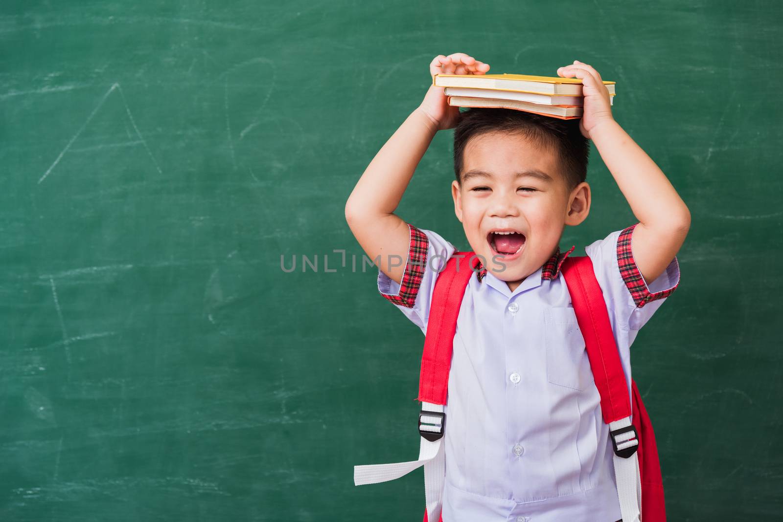 Back to School. Happy Asian funny cute little child boy from kindergarten in student uniform with school bag and book on head smiling on green school blackboard, First time to school education concept